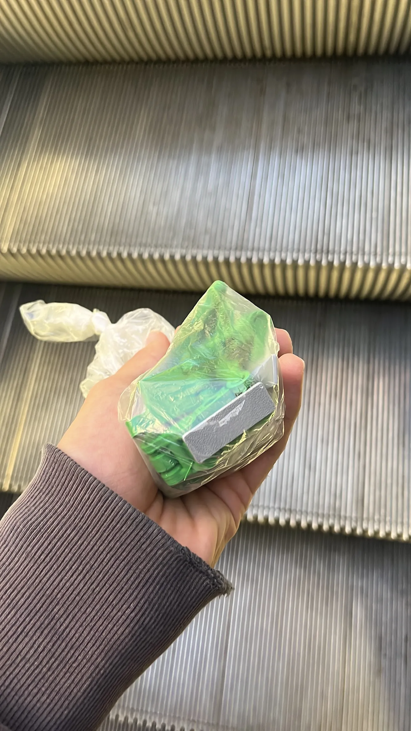 A hand holding a small bag of green 3D-printed pieces above an escalator.